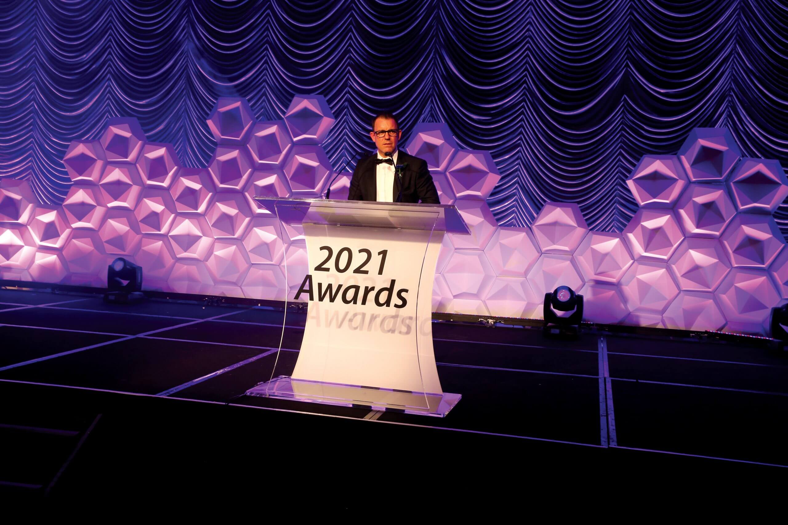 A person in formal attire speaks at a podium labeled "2021 Awards" on a stage with a geometric and draped backdrop, offering insight about us.