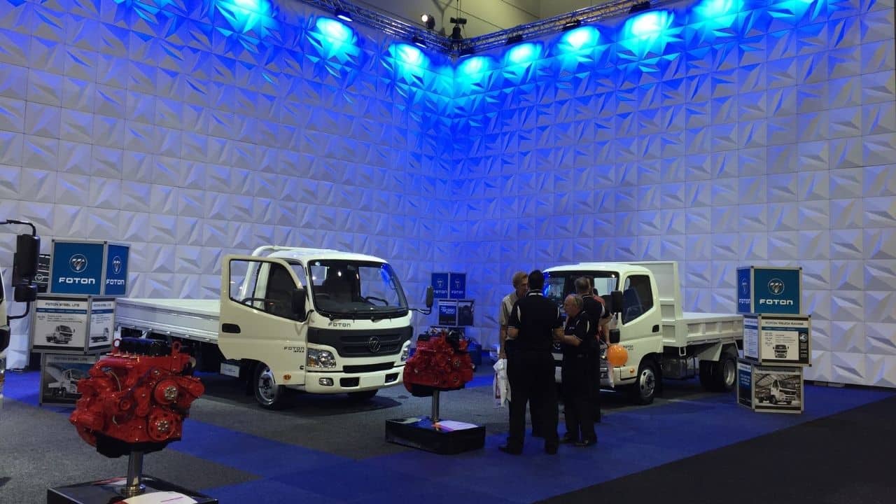 Trade show with stealth wall backdrop and blue lighting. 2 white trucks and two red vehicle engines are displayed in front of the wall.