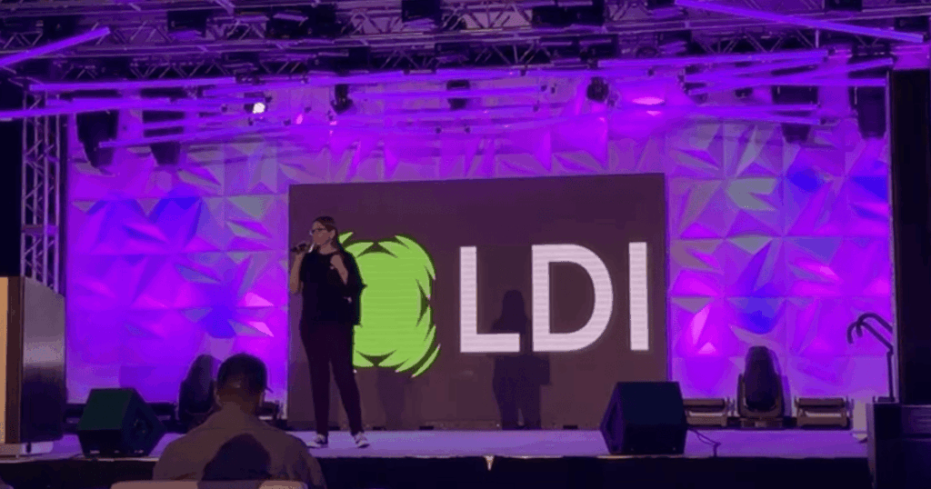 A person stands on a stage with a microphone in front of a large screen displaying the letters "LDI" and a green graphic, under purple lighting, at an event highlighting the mod scenes partnership.
